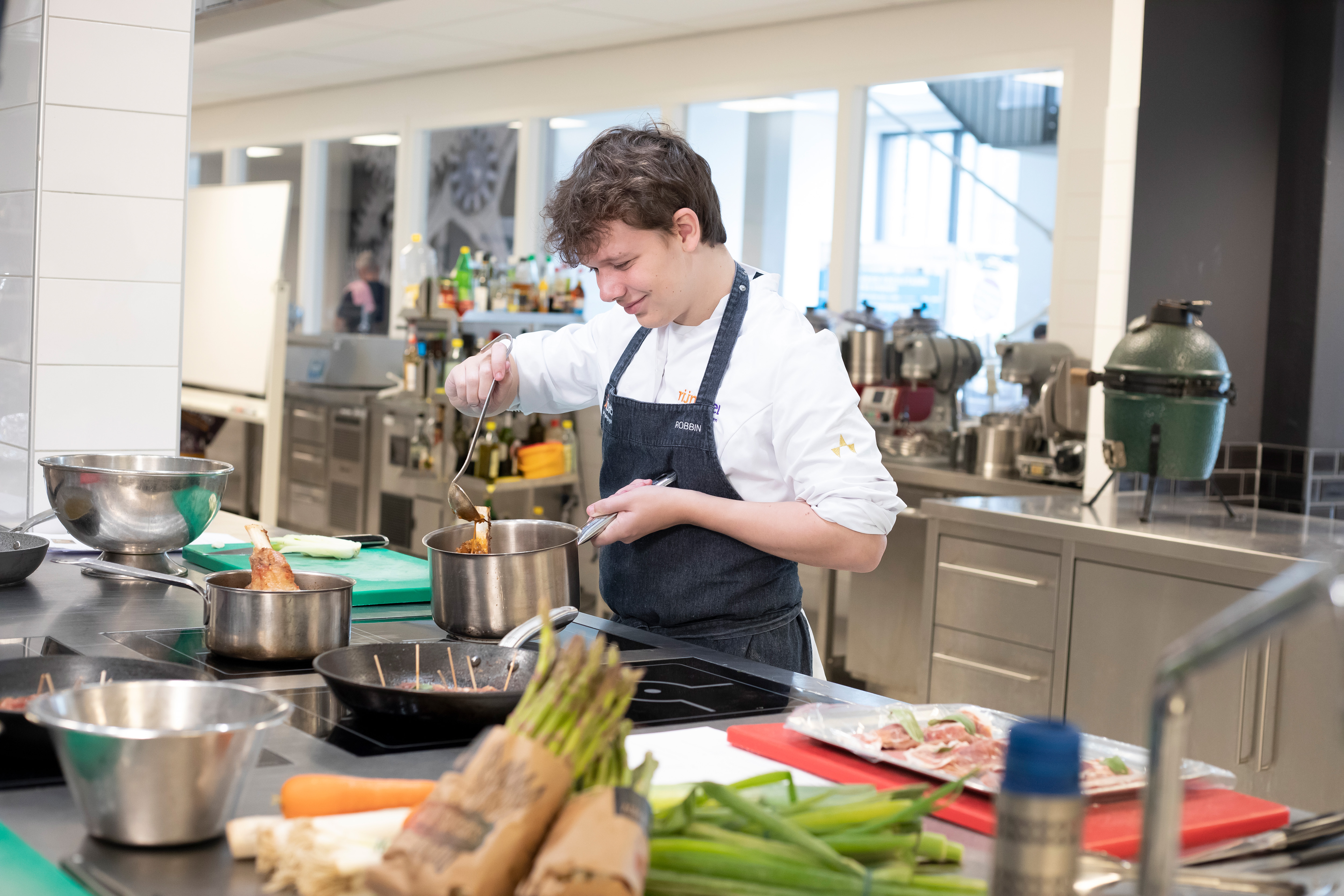 Jongen in kokskleding is aan het koken met een steelpan en heeft een lepel in zijn hand