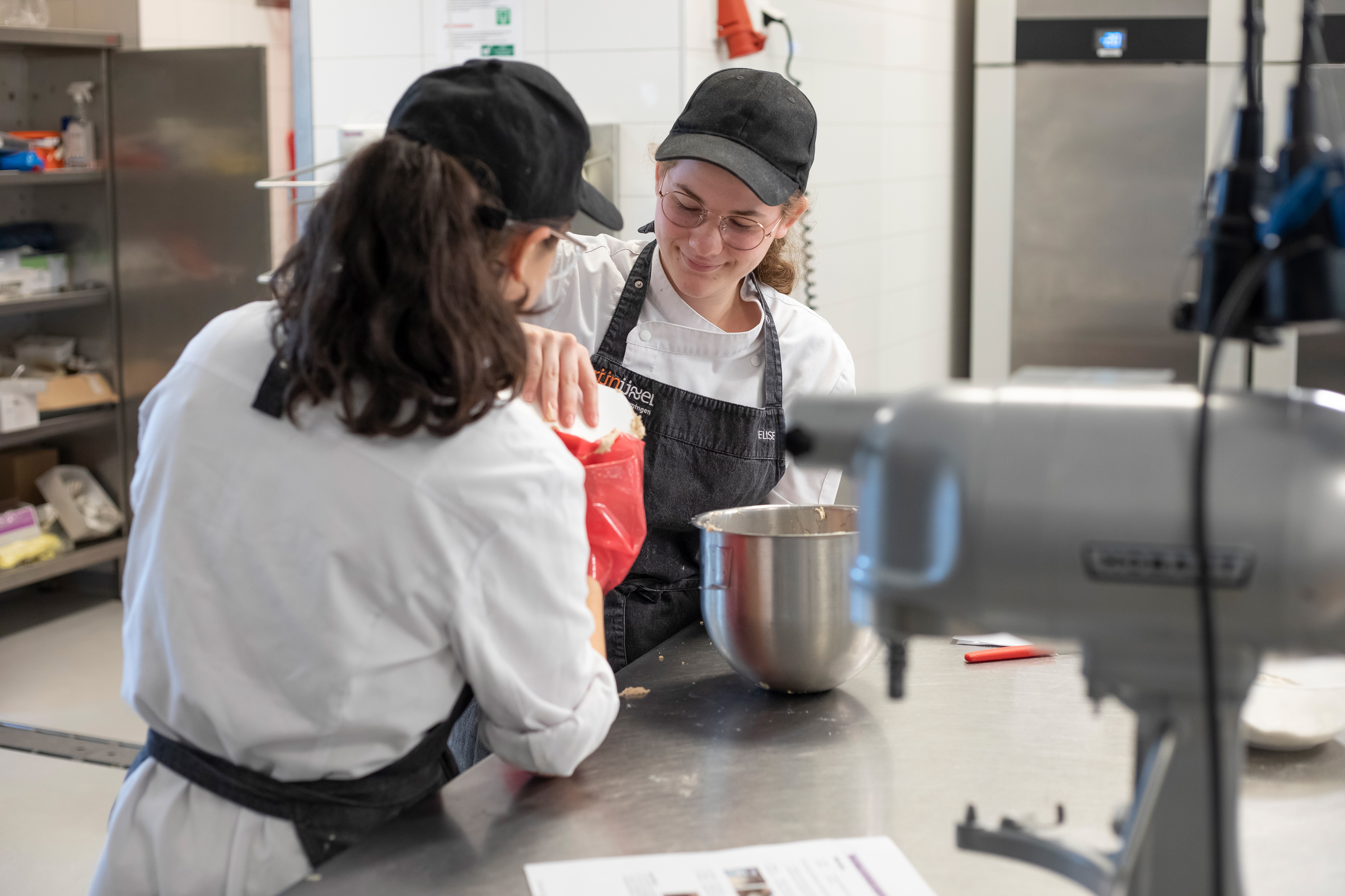 Twee meisjes in bakkerskleding staan bij een mixer en doen beslag in een spuitzak
