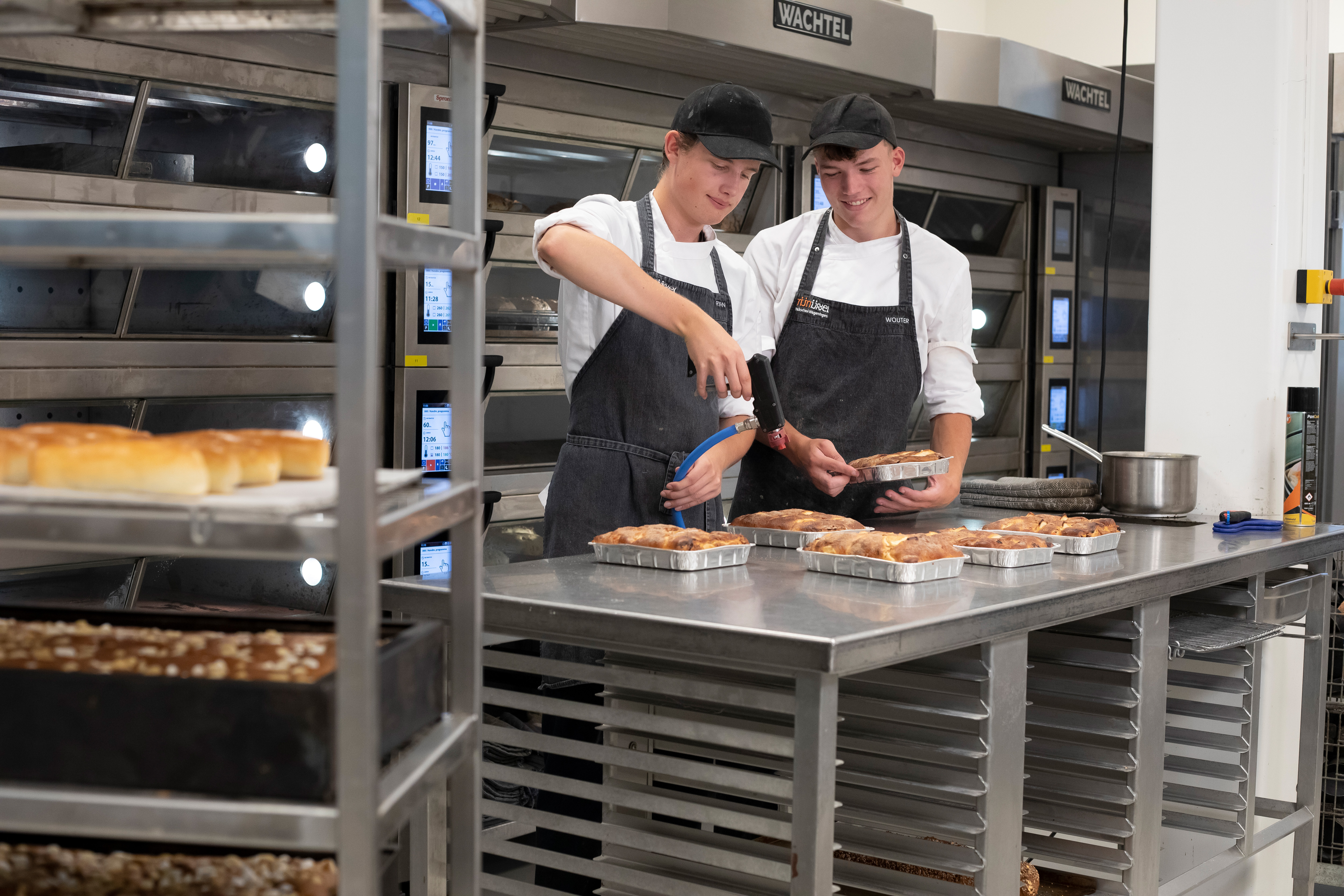 Je ziet twee jongens in de bakkerij met bakkerskleding aan. Op de voorgrond zie je een rek met broden en op de achtergrond zie je de jongens broodgerechten bewerken met een brander