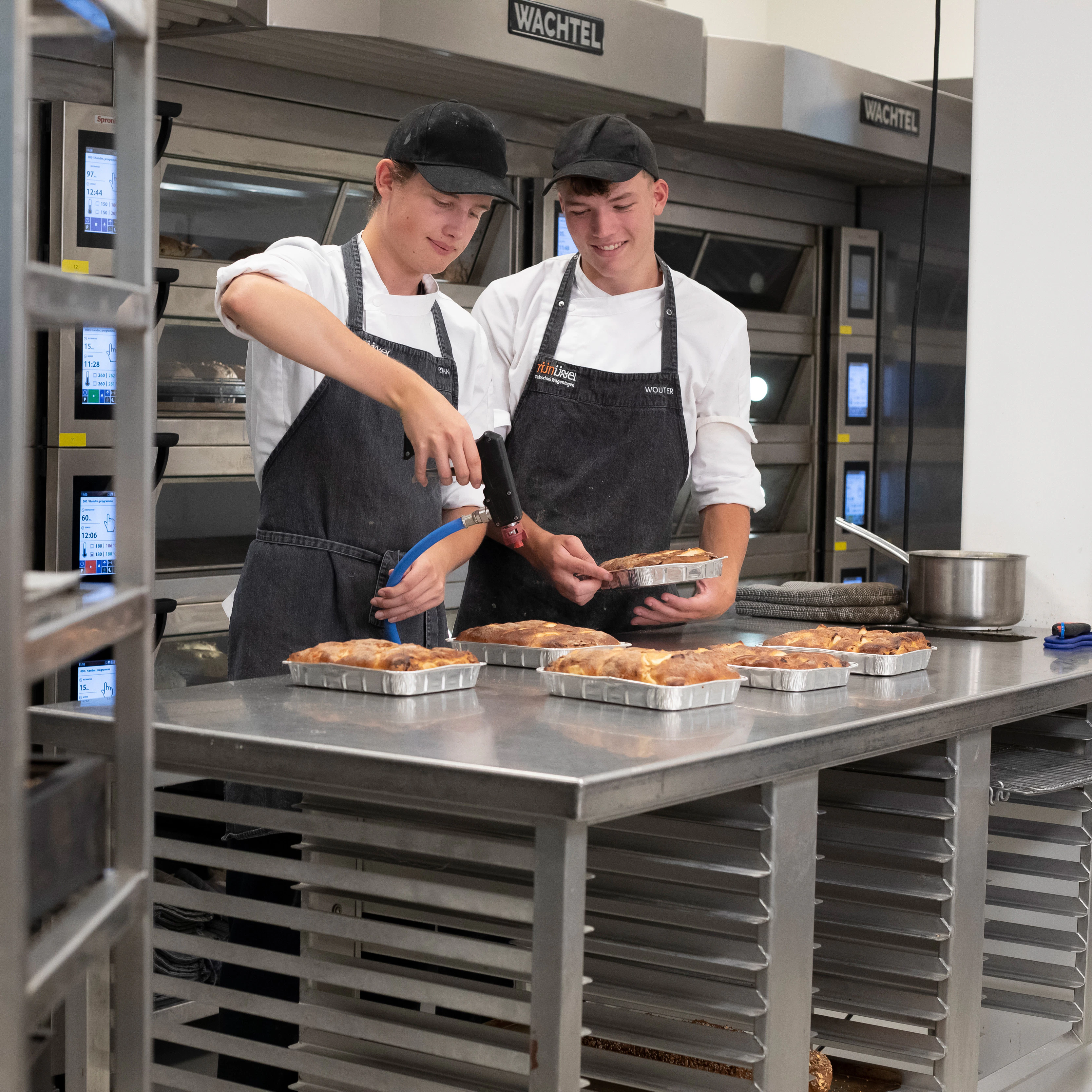 Je ziet twee jongens in de bakkerij met bakkerskleding aan. Op de voorgrond zie je een rek met broden en op de achtergrond zie je de jongens broodgerechten bewerken met een brander