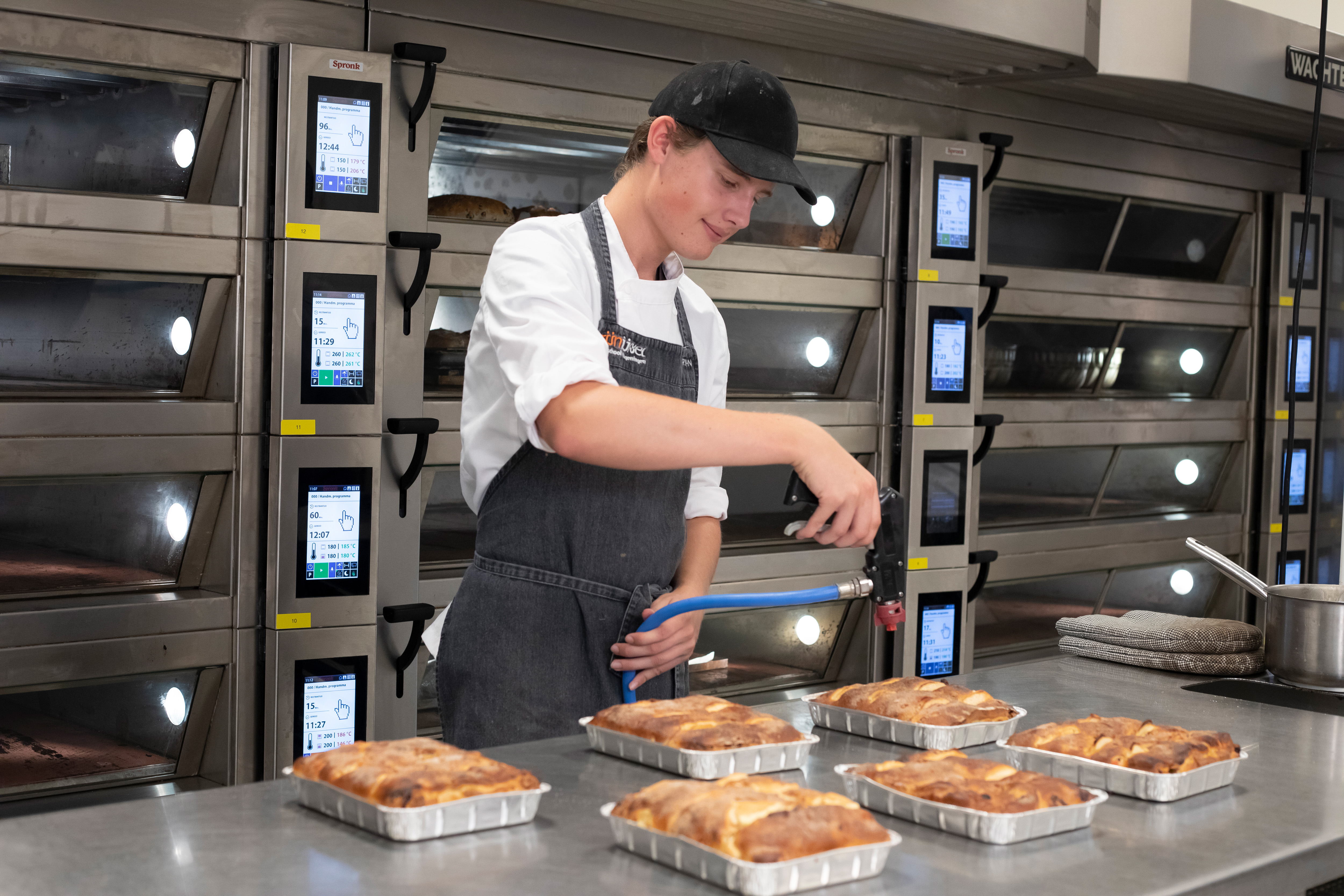Jongen staat in bakkerskleding in de bakkerij met zes broodgerechten voor hem die hij aan het bewerken is met een brander