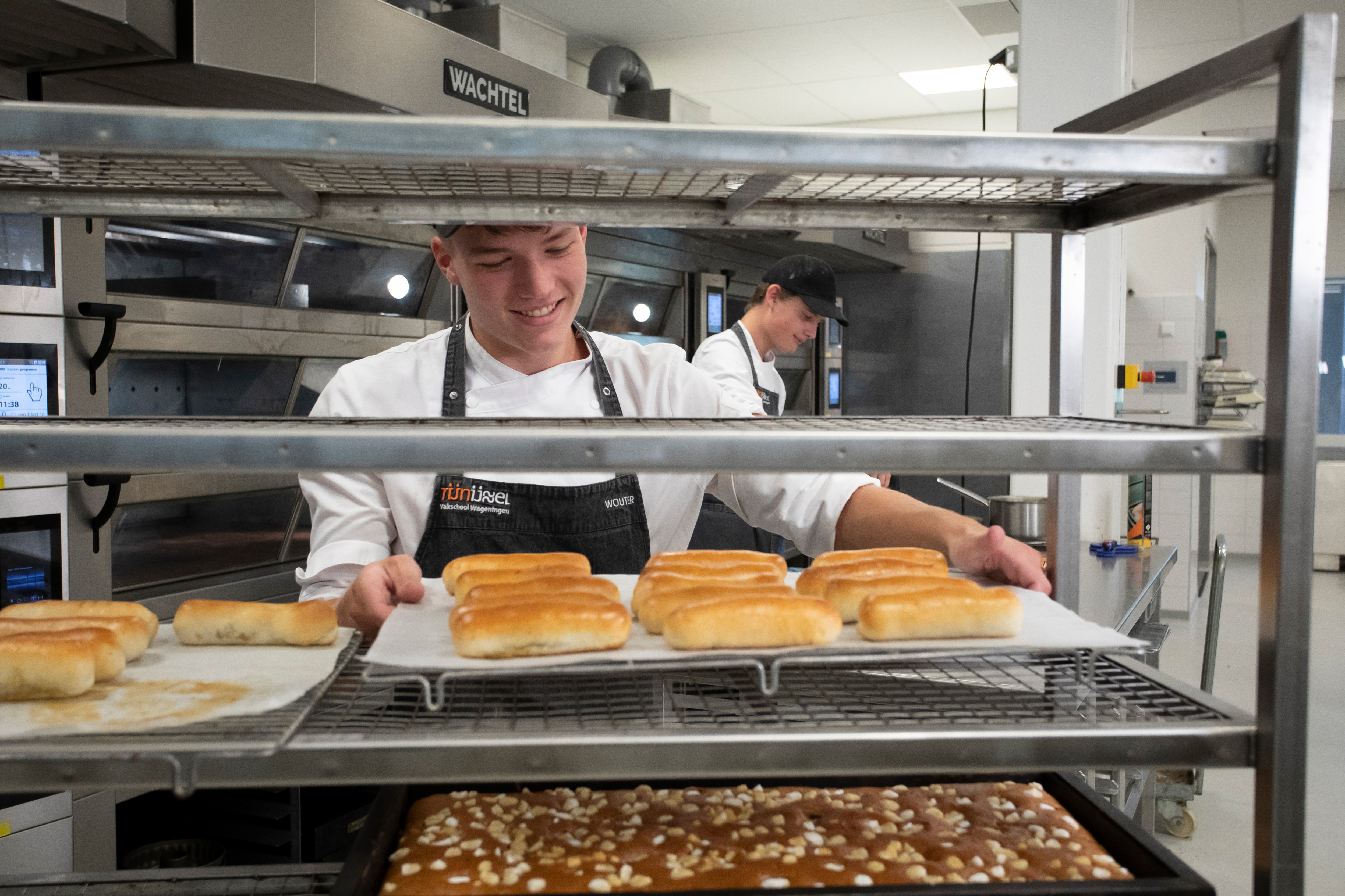 Jongen in bakkerskleding in een bakkerij staat achter een rek waar broden en cakejes liggen