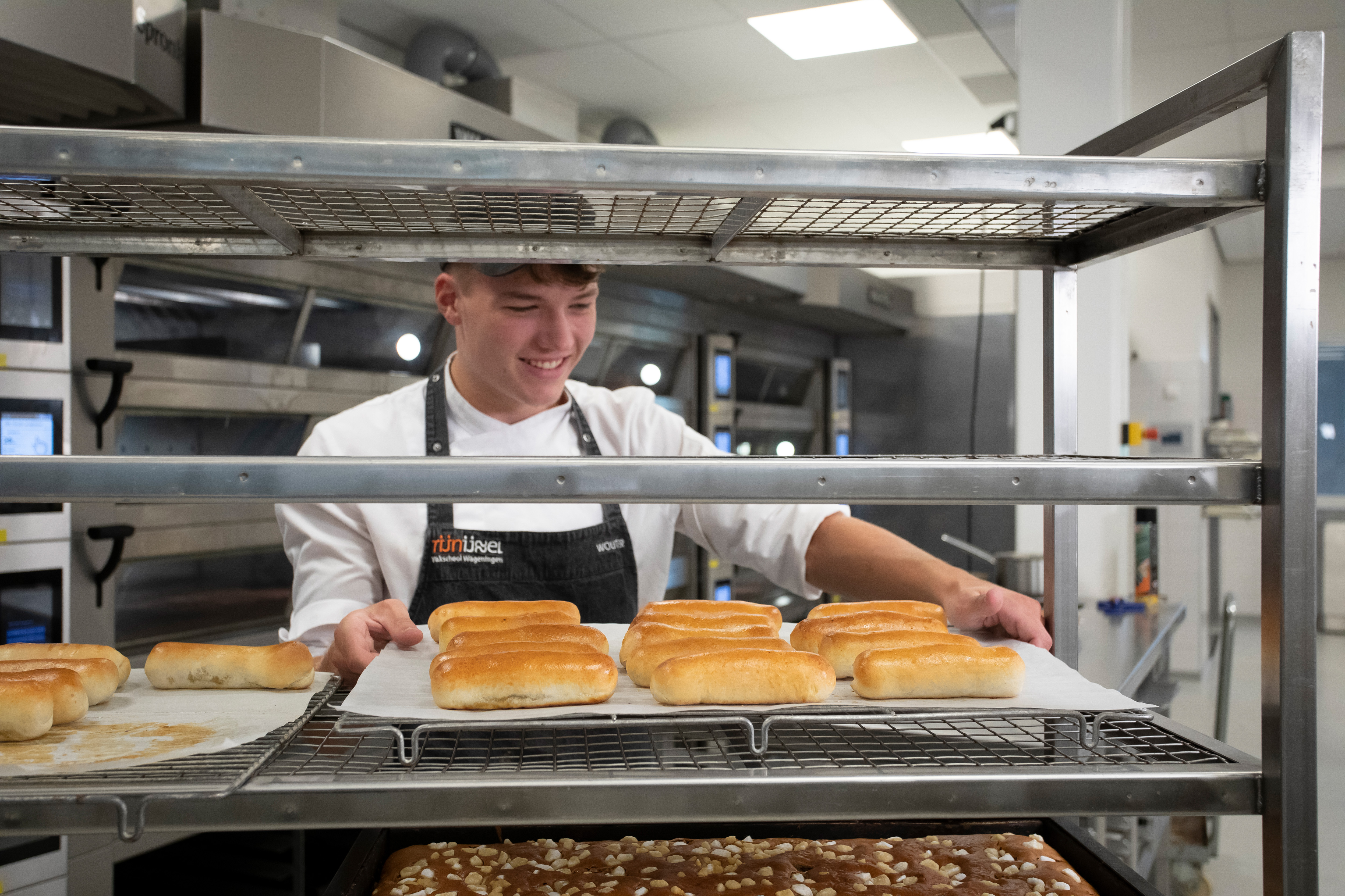 Jongen in bakkerskleding in een bakkerij staat achter een rek waar broden en cakejes liggen