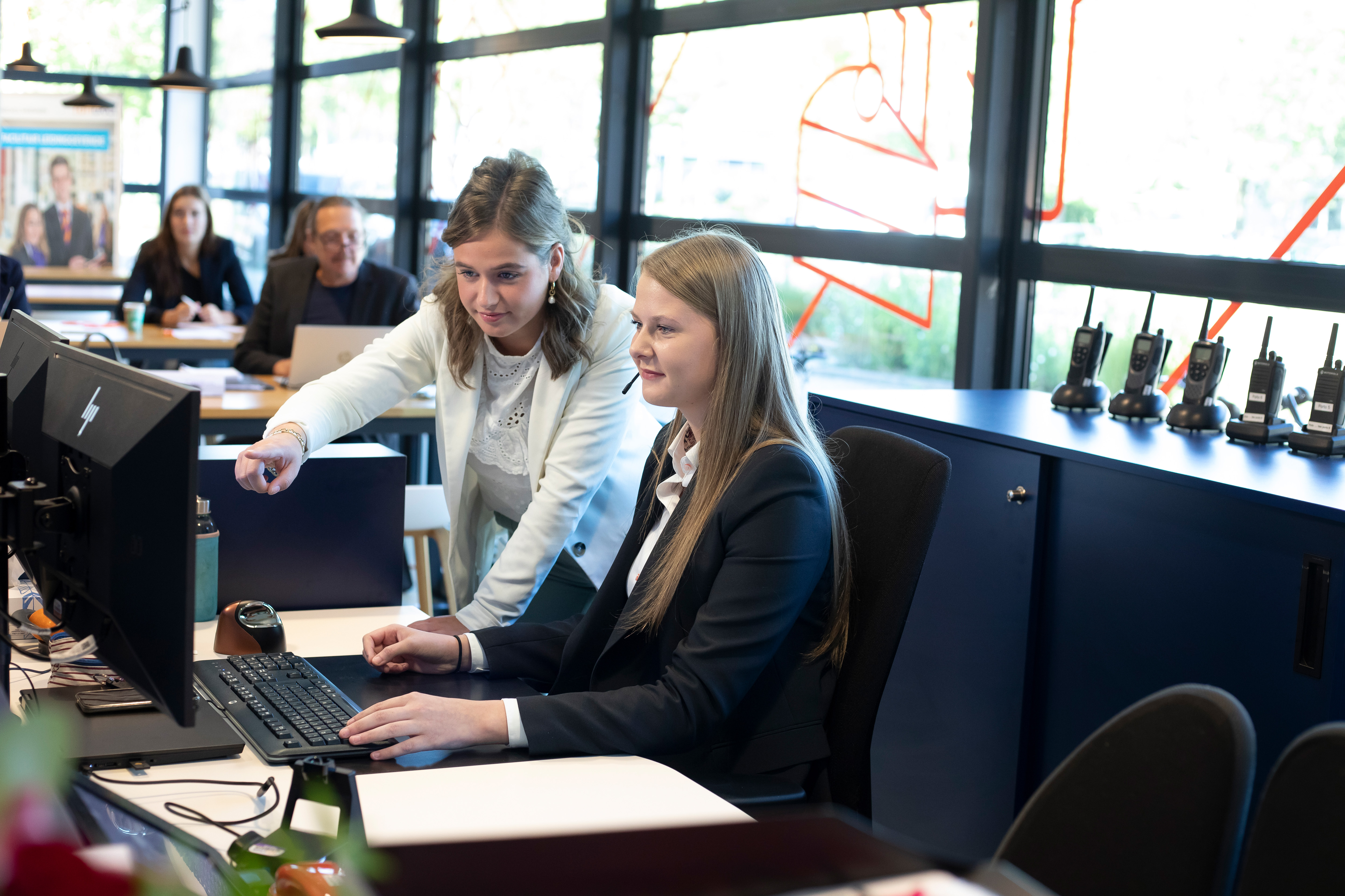 Een meisje in een net pak zit achter de computer terwijl er naast een een ander meisje naar het beeldscherm wijst en haar iets laat zien