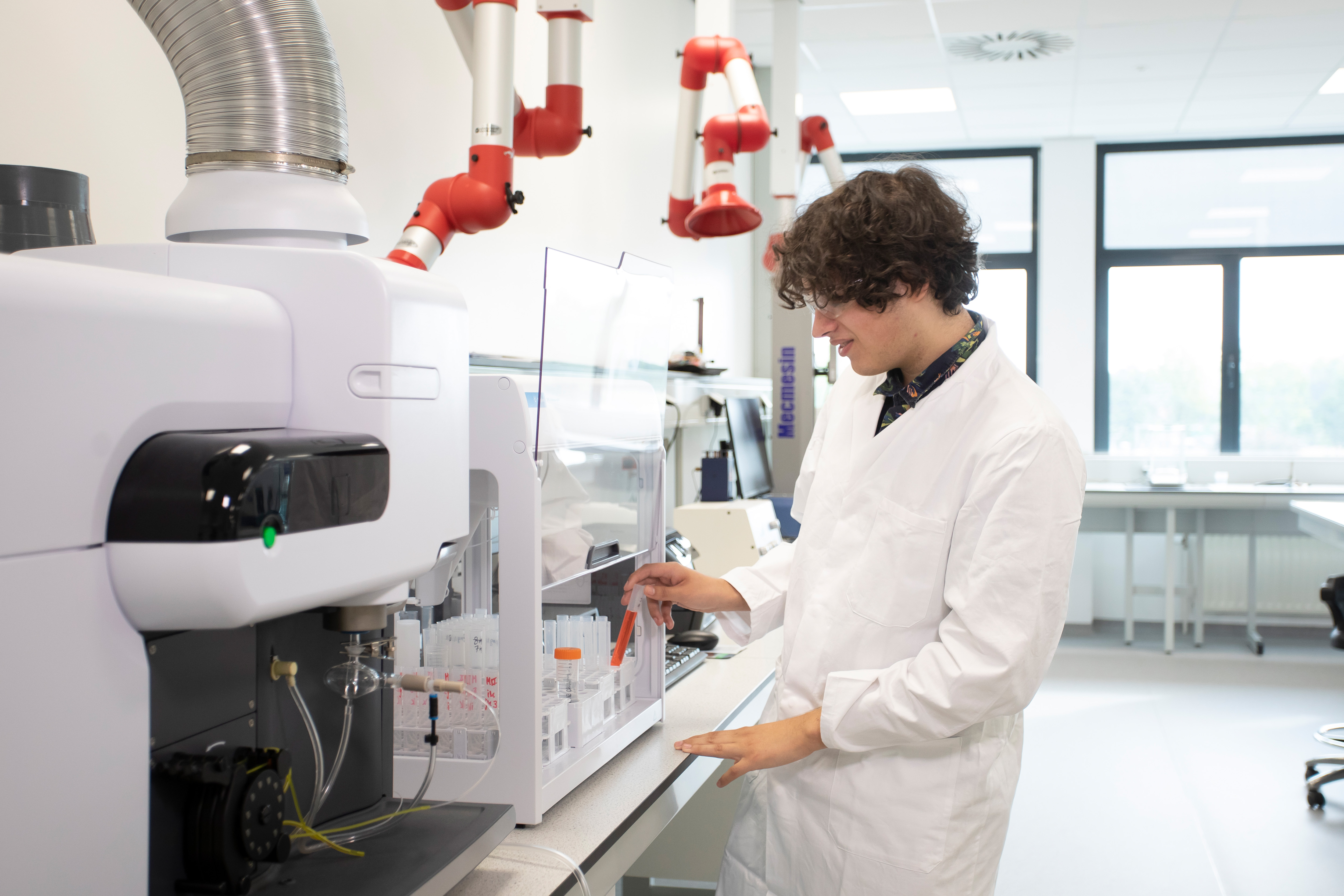 Jongen in een witte labaratoriumjas is bezig bij een machine en heeft oranje chemische vloeistoffen in buisjes