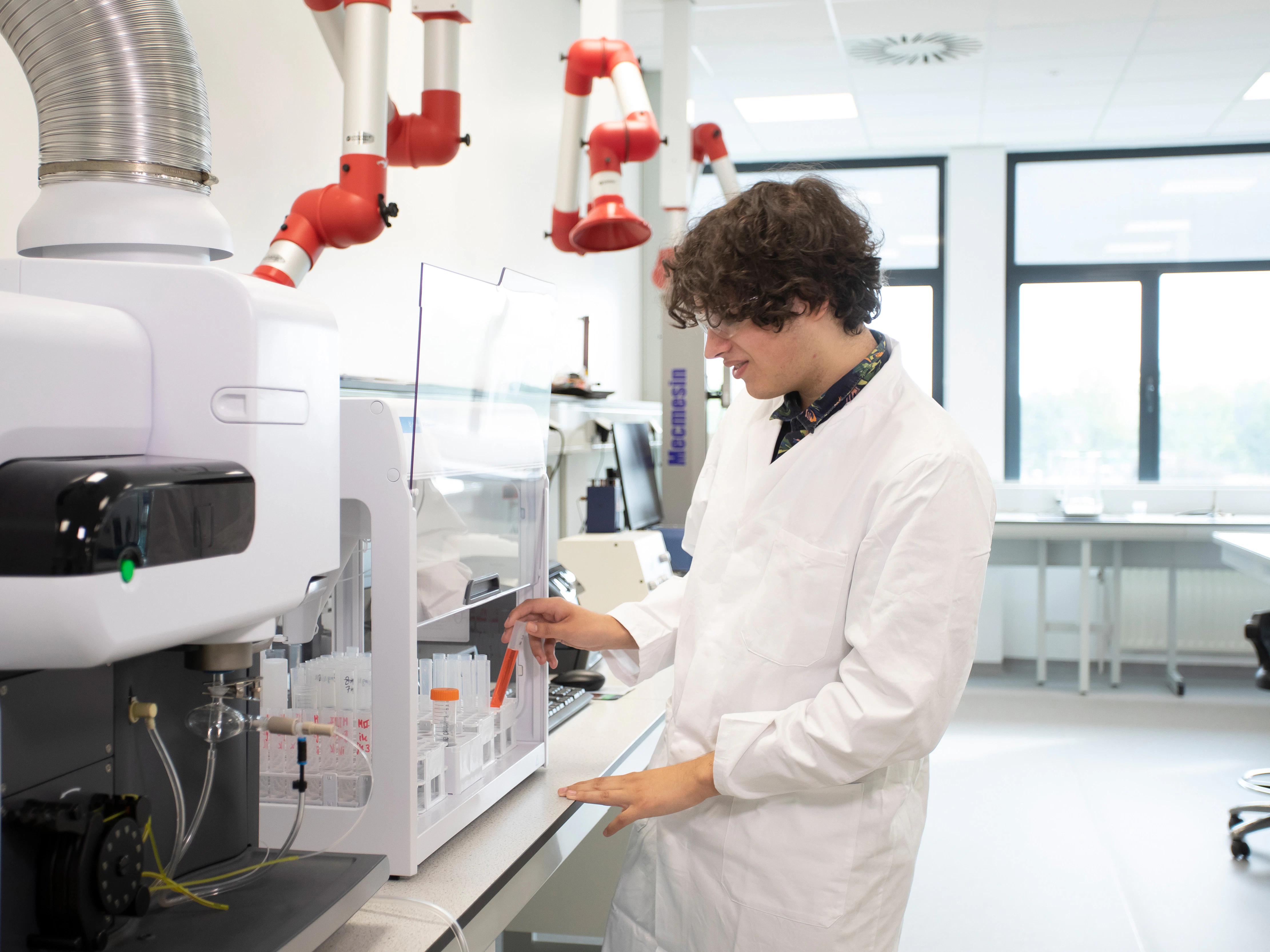 Jongen in een witte labaratoriumjas is bezig bij een machine en heeft oranje chemische vloeistoffen in buisjes