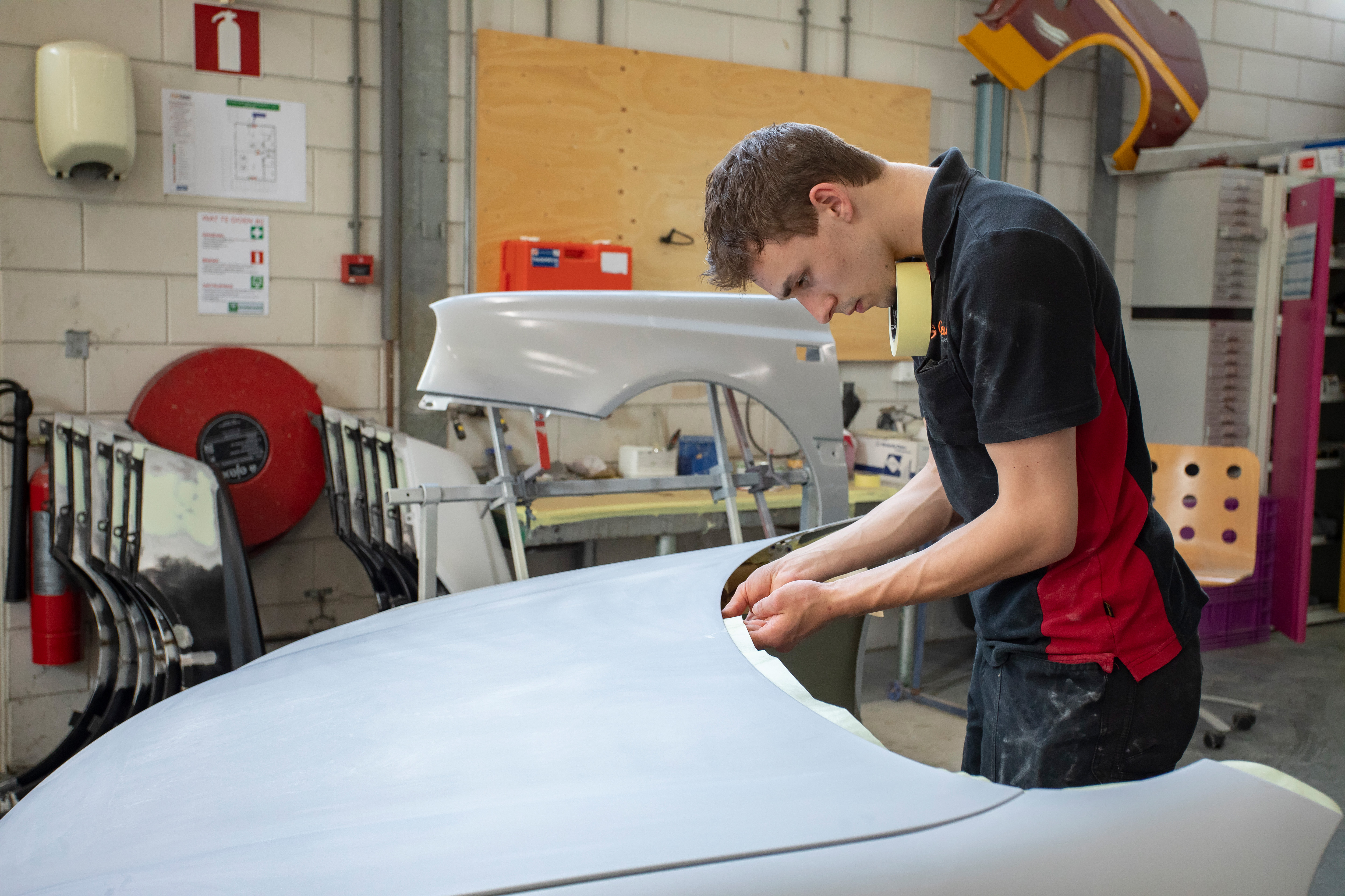 Jongen in werkkleding plakt tape op een onderdeel van de auto terwijl hij in de werkplaats staat