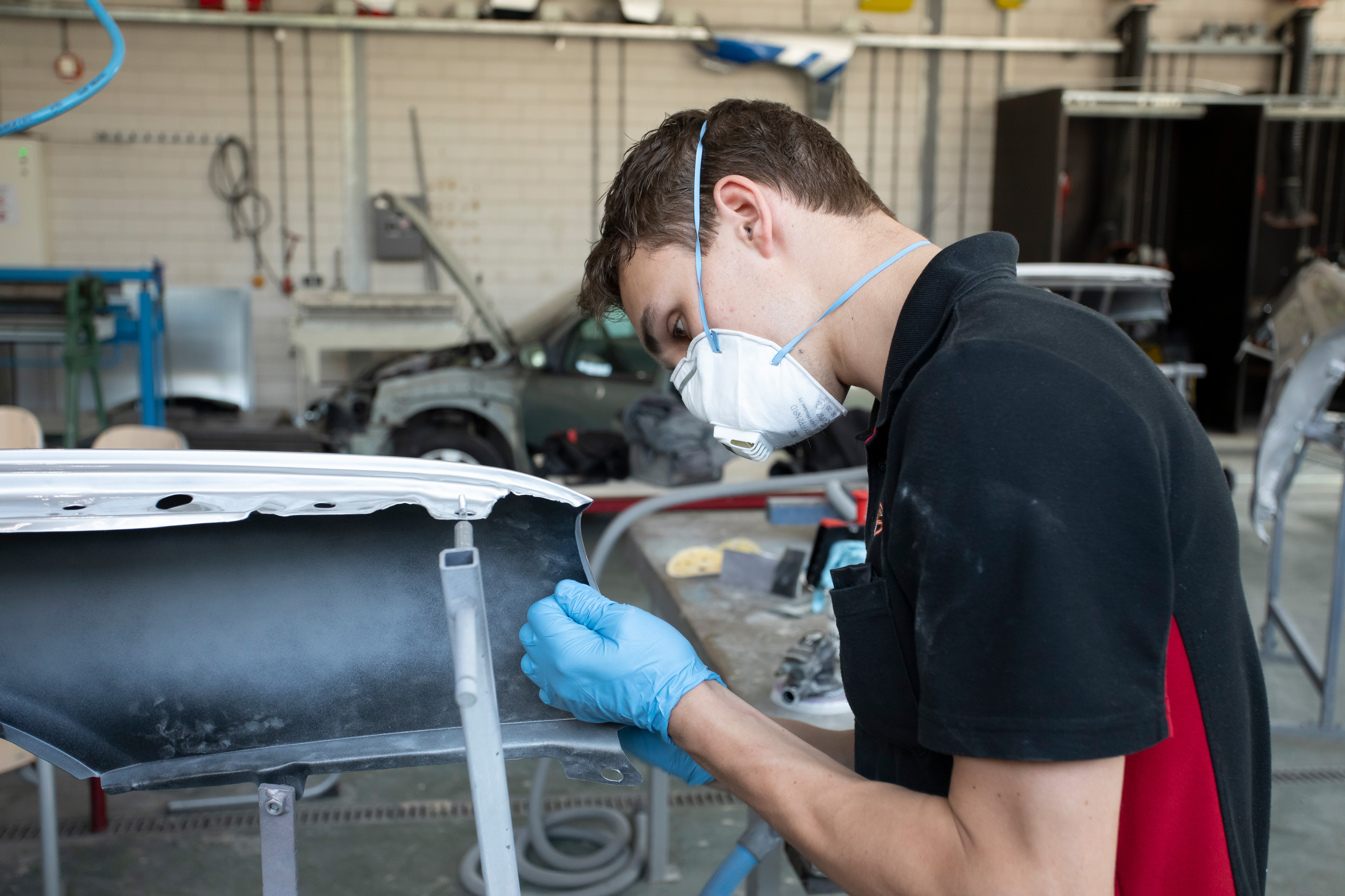 Jongen met een mondkapje op staat in de werkplaats en is bezig met een onderdeel van de auto