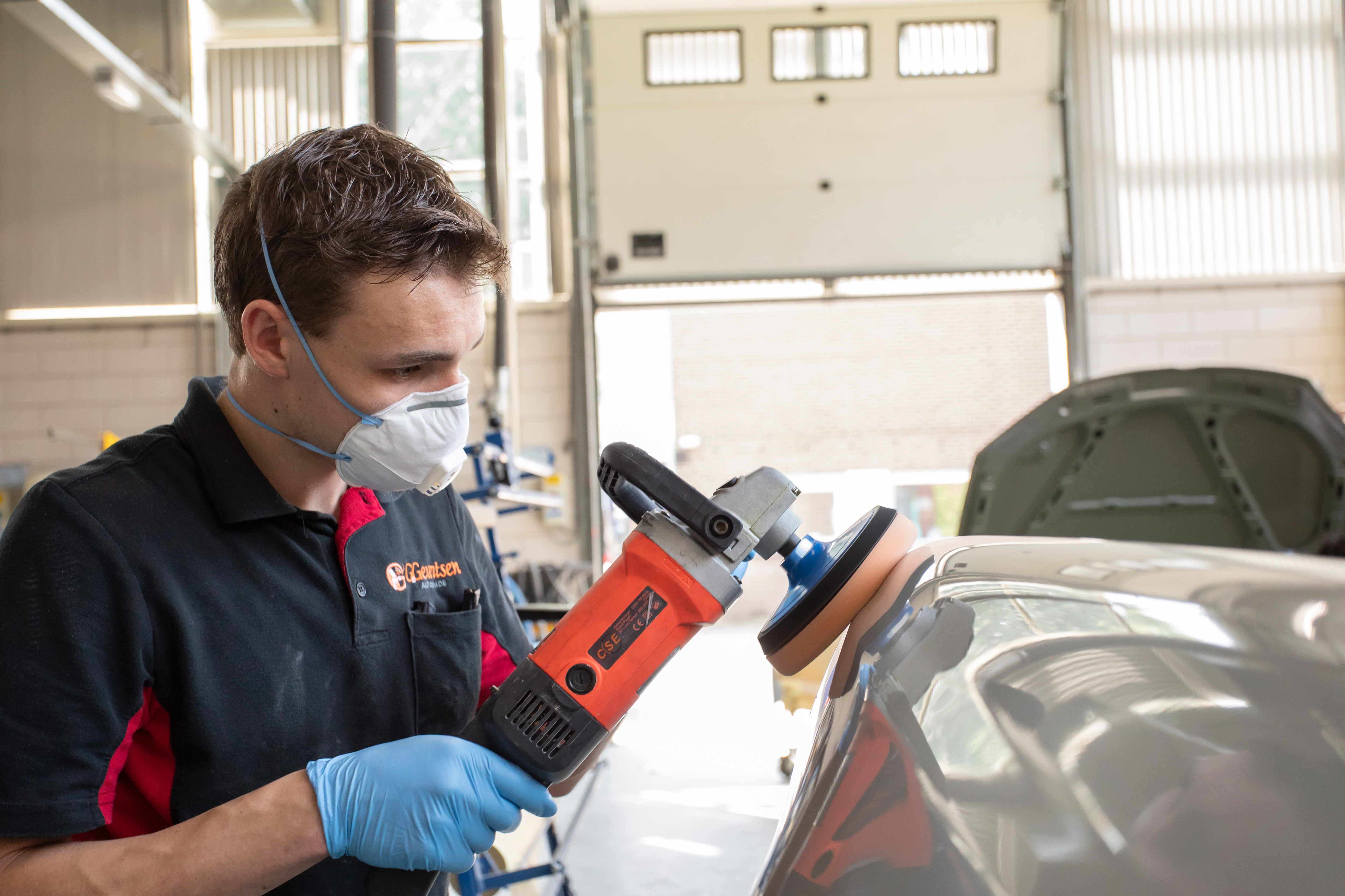 Jongen met een mondkapje op heeft een apparaat in de hand waarmee hij een onderdeel van de auto aan het boenen is