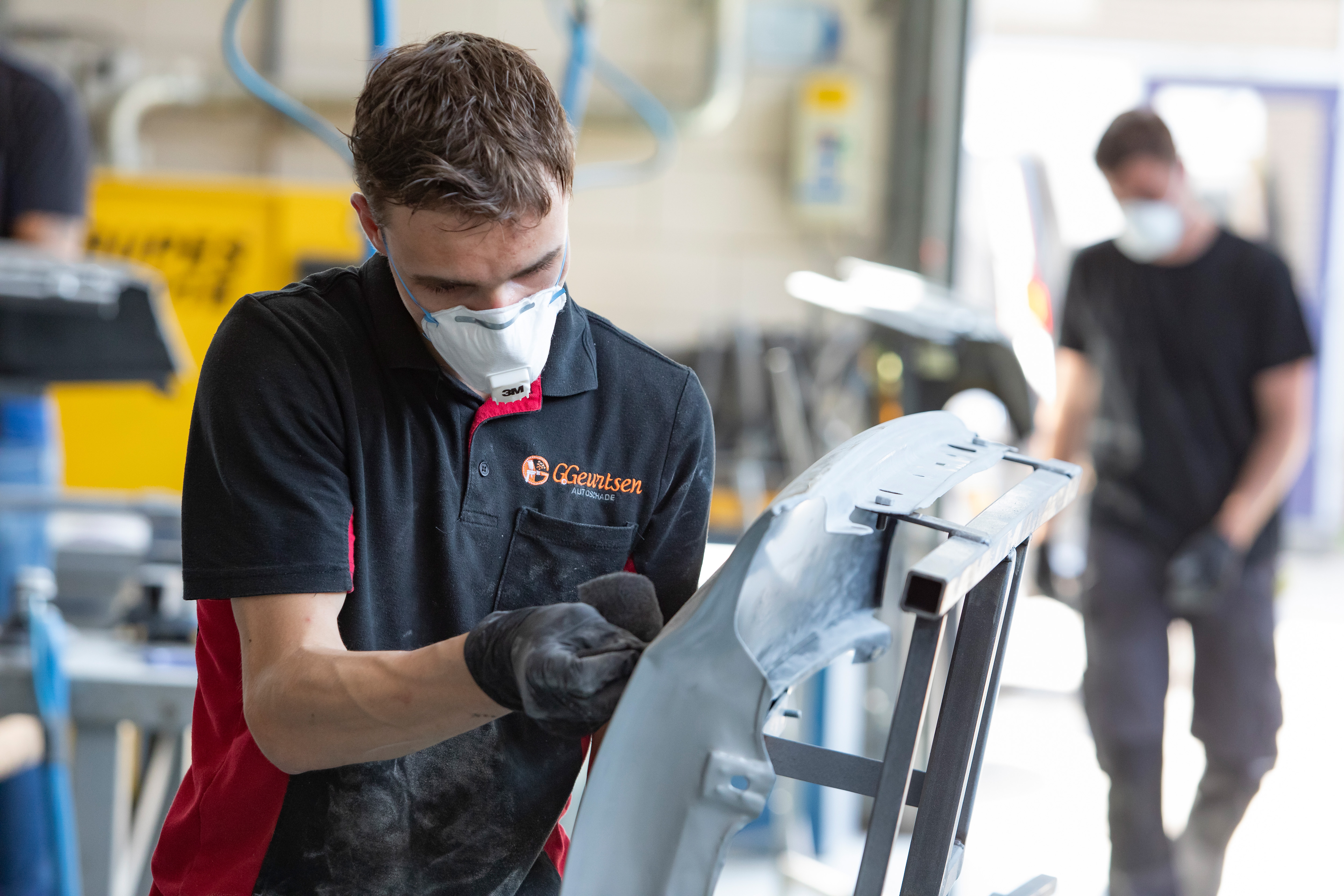 Jongen met een mondkapje op en werkkleding aan is een onderdeel van de auto aan het schuren met schuurpapier